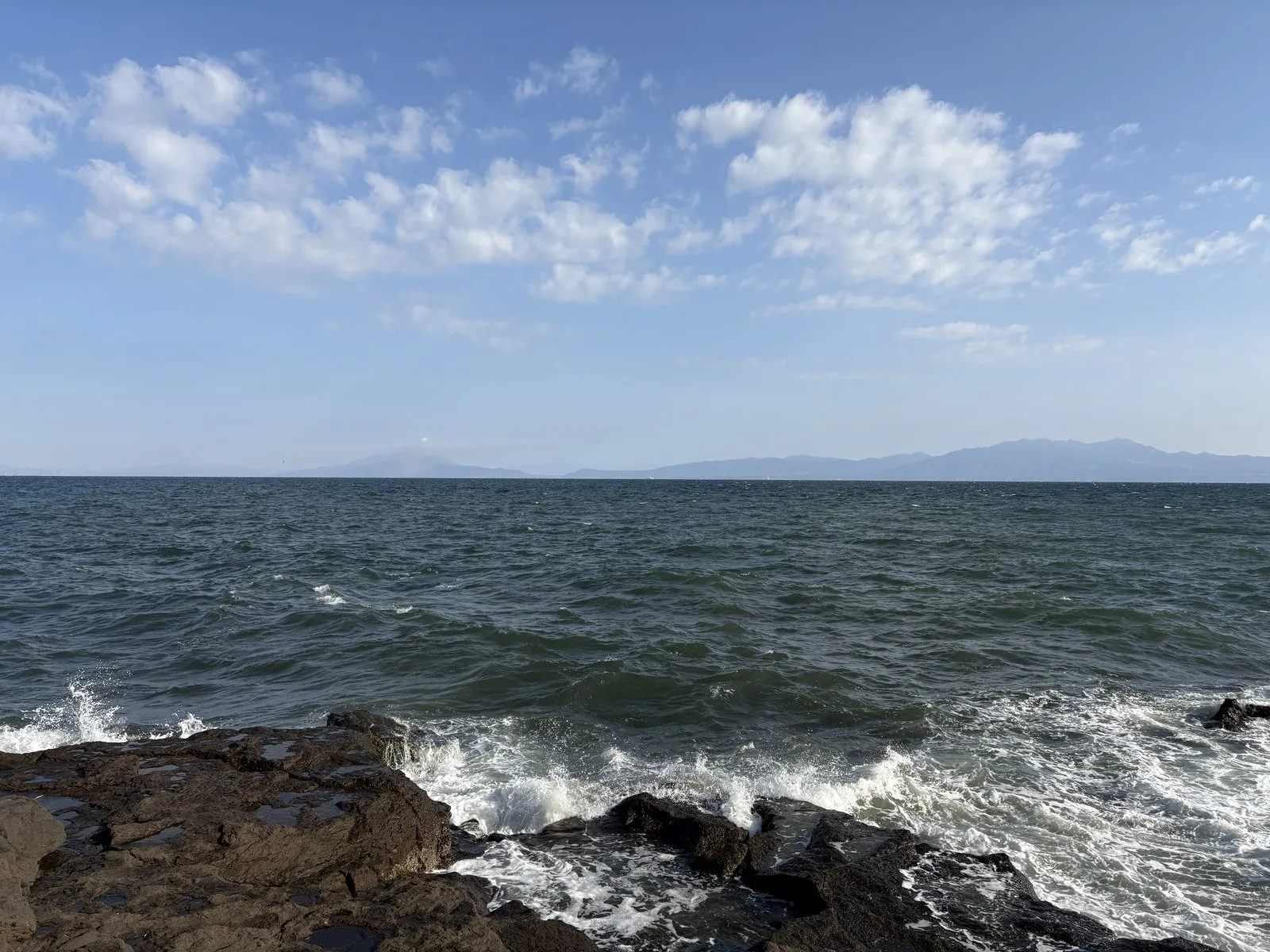 道の駅いぶすきの下の黒い岩場に波が打ち寄せる。錦江湾の対岸に大隅半島が霞む