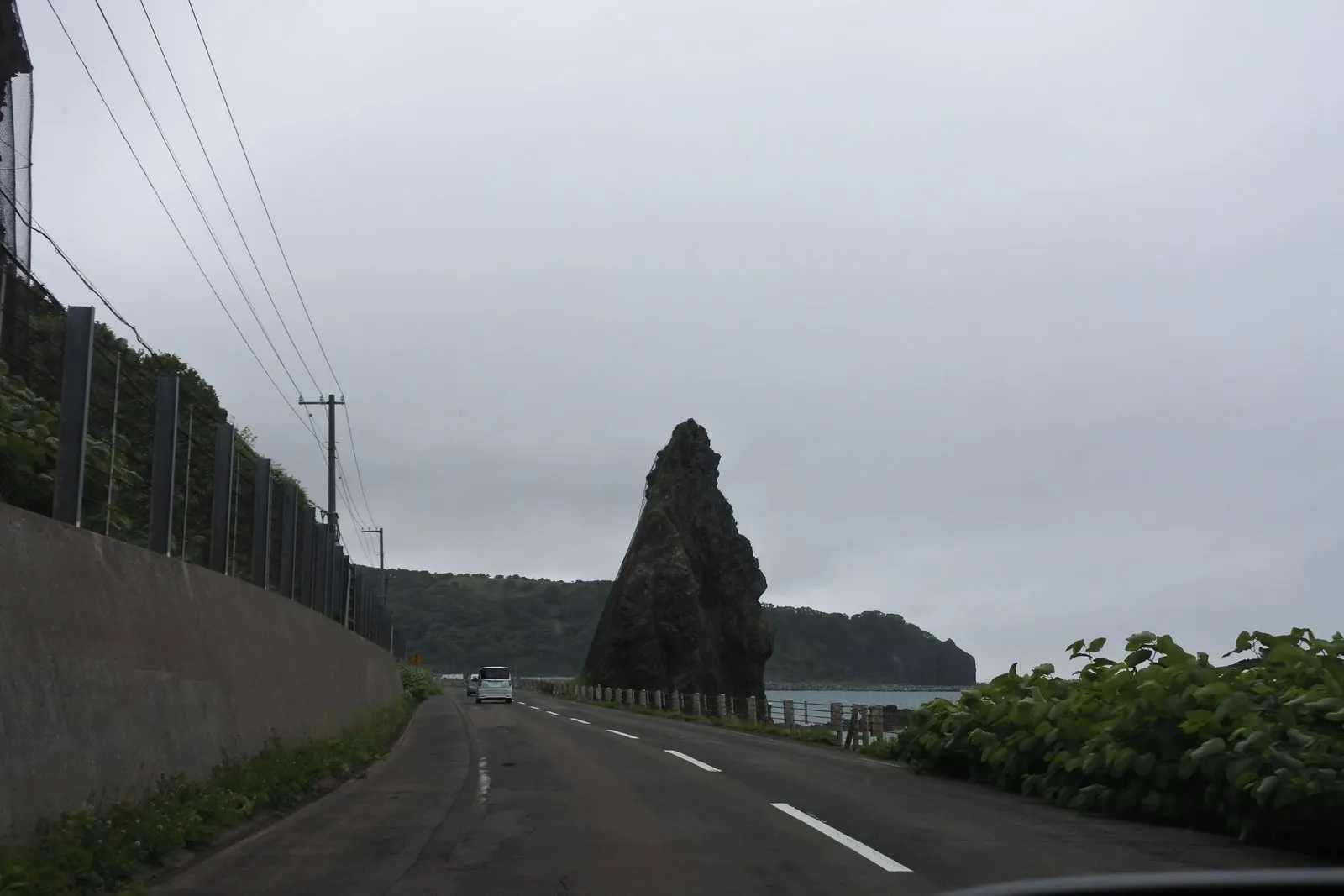 国道の先、海沿いにそそり立つ黒い三角形の奇岩。空は曇り、路面はまだ濡れている