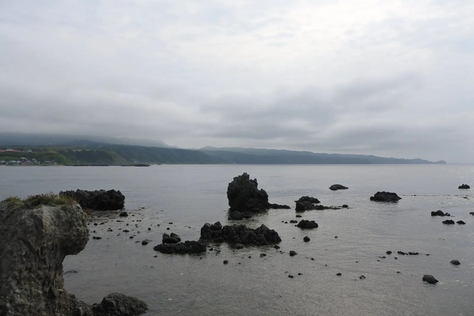 海面から突き出した黒い岩礁が点々と並ぶ海岸。奥に対岸の陸地と集落が見える